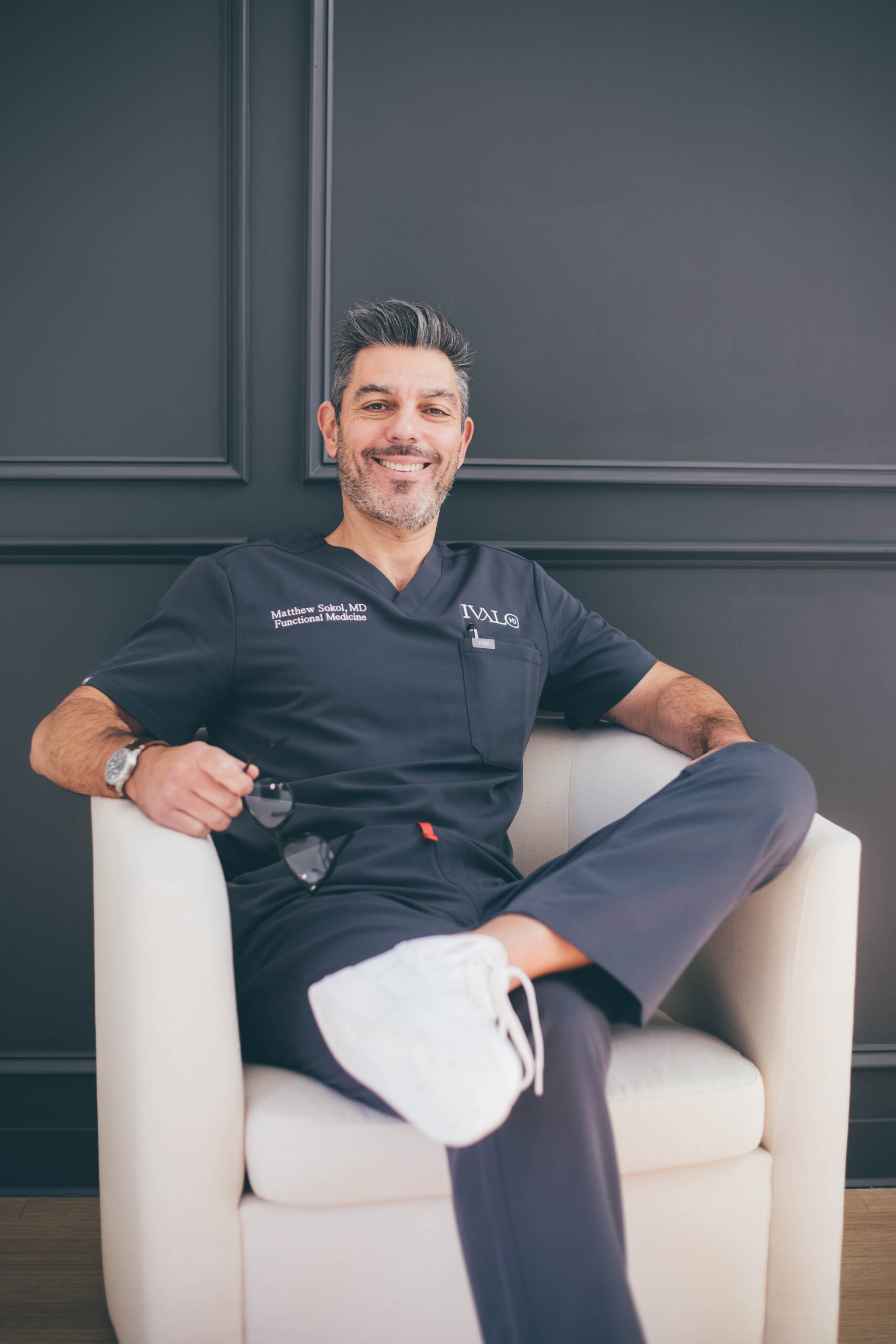 The doctor sitting to discuss functional medicine in Dallas. Dr. Matthew Sokol sitting in a white chair against a dark wall, wearing navy blue medical scrubs.
