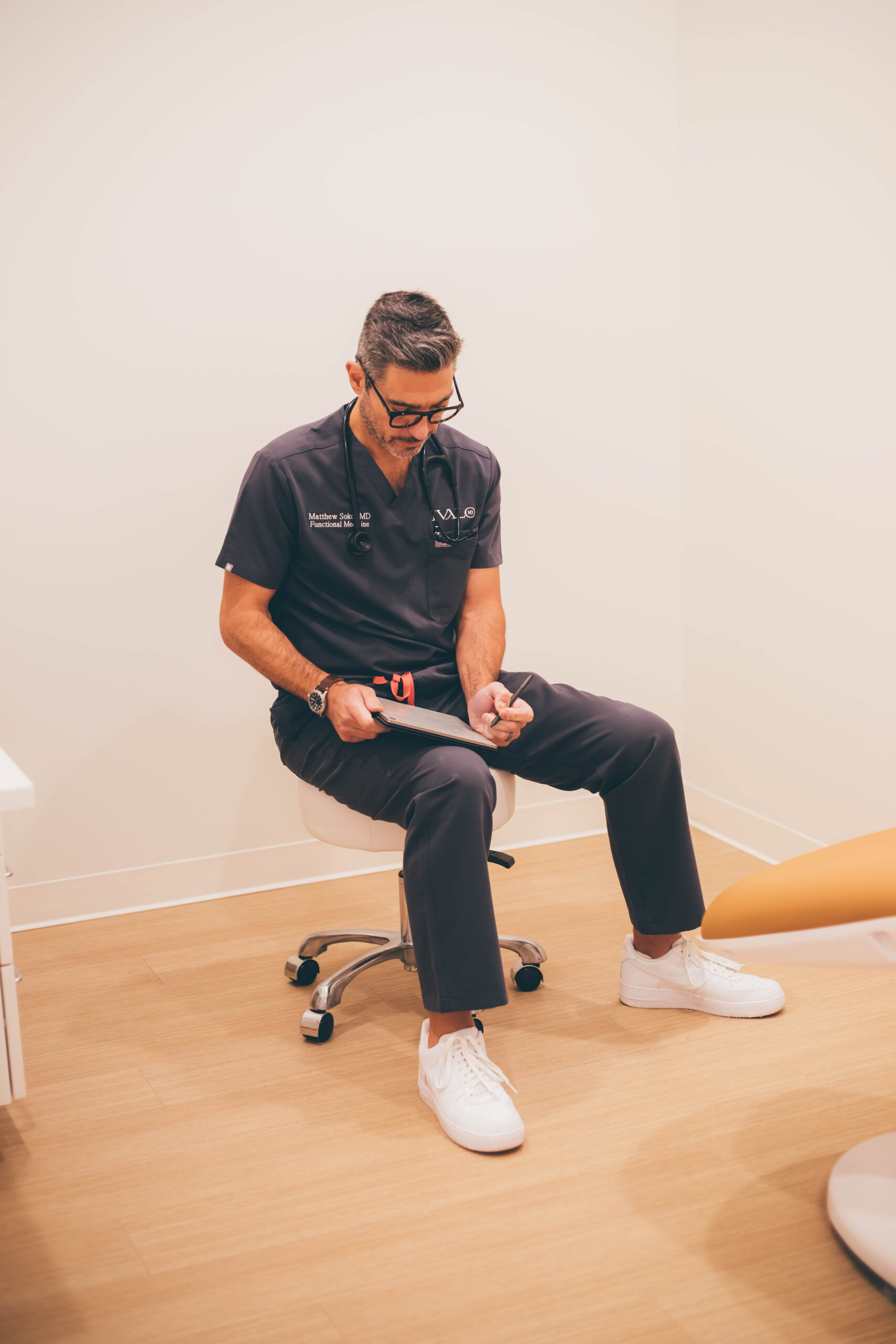 Dr. Sokol sitting on a stool in a hallway, wearing navy scrubs and holding a tablet.